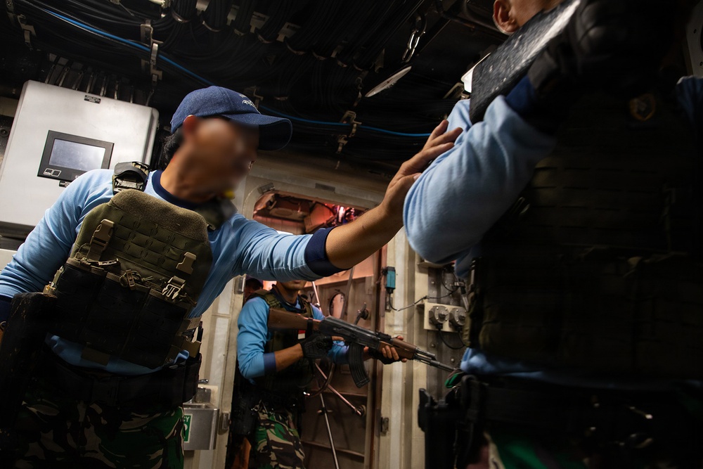 The image shows what appears to be a military or law enforcement boarding/clearance drill taking place inside a ship or vessel. Personnel wearing tactical vests, blue shirts, and camouflage pants are conducting a room-clearing exercise in a confined interior space. At least one individual is carrying what appears to be an AK-pattern rifle.
