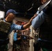 The image shows what appears to be a military or law enforcement boarding/clearance drill taking place inside a ship or vessel. Personnel wearing tactical vests, blue shirts, and camouflage pants are conducting a room-clearing exercise in a confined interior space. At least one individual is carrying what appears to be an AK-pattern rifle.