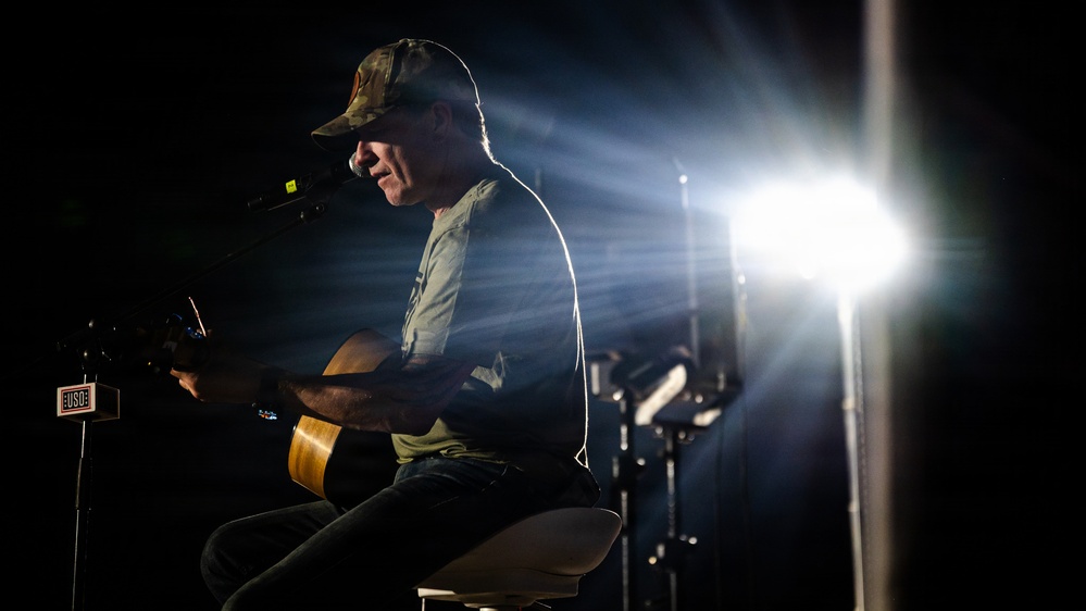 A male performer sits on a stool playing an acoustic guitar in front of a microphone on a darkened stage. The scene is dramatically lit by a bright stage light from behind and to the right, creating strong rim lighting and lens flare effects. A small USO logo sign is visible on a stand to the left, indicating this is a USO performance.