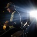 A male performer sits on a stool playing an acoustic guitar in front of a microphone on a darkened stage. The scene is dramatically lit by a bright stage light from behind and to the right, creating strong rim lighting and lens flare effects. A small USO logo sign is visible on a stand to the left, indicating this is a USO performance.