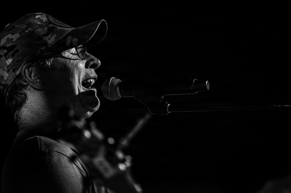 A black and white photograph of a male performer singing into a microphone on stage. He is wearing a camouflage baseball cap and glasses, captured in dramatic side-profile lighting against a dark background. A guitar neck is partially visible in the foreground, suggesting a live music performance.