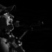 A black and white photograph of a male performer singing into a microphone on stage. He is wearing a camouflage baseball cap and glasses, captured in dramatic side-profile lighting against a dark background. A guitar neck is partially visible in the foreground, suggesting a live music performance.