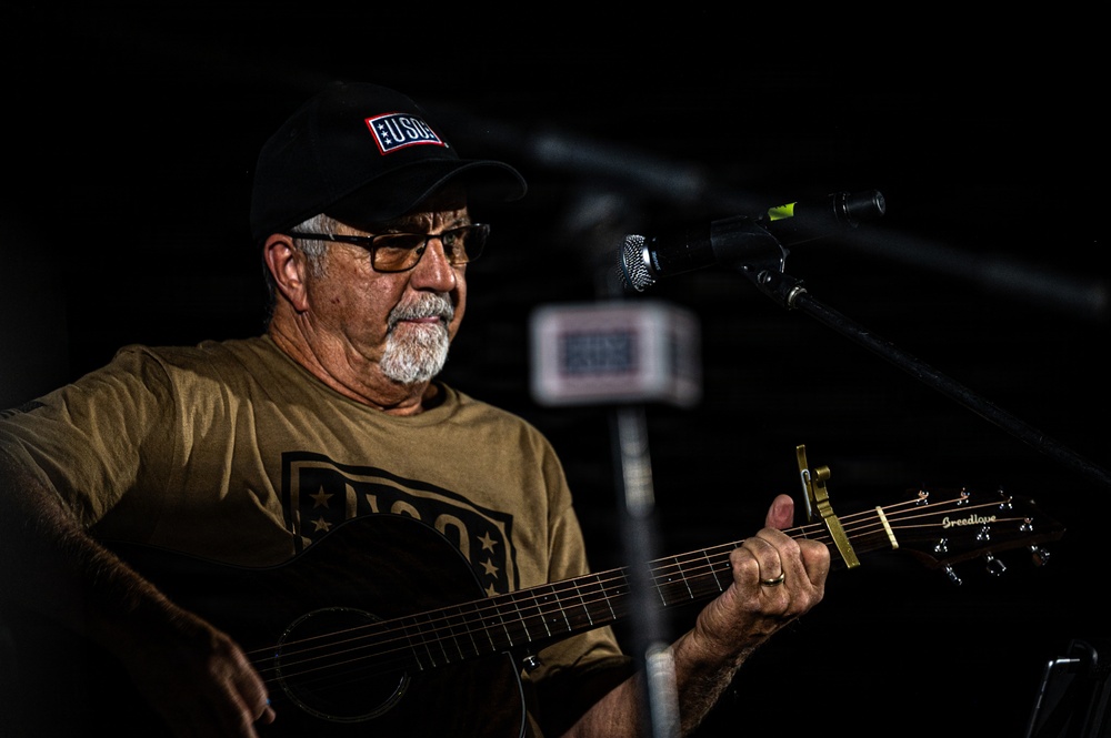 An older man is performing live music on an acoustic guitar in a dimly lit venue. He is wearing USO-branded clothing including a black cap and olive green t-shirt, suggesting a USO entertainment event. A microphone on a stand is positioned in front of him, and a gold capo is visible on the guitar neck.