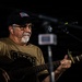 An older man is performing live music on an acoustic guitar in a dimly lit venue. He is wearing USO-branded clothing including a black cap and olive green t-shirt, suggesting a USO entertainment event. A microphone on a stand is positioned in front of him, and a gold capo is visible on the guitar neck.