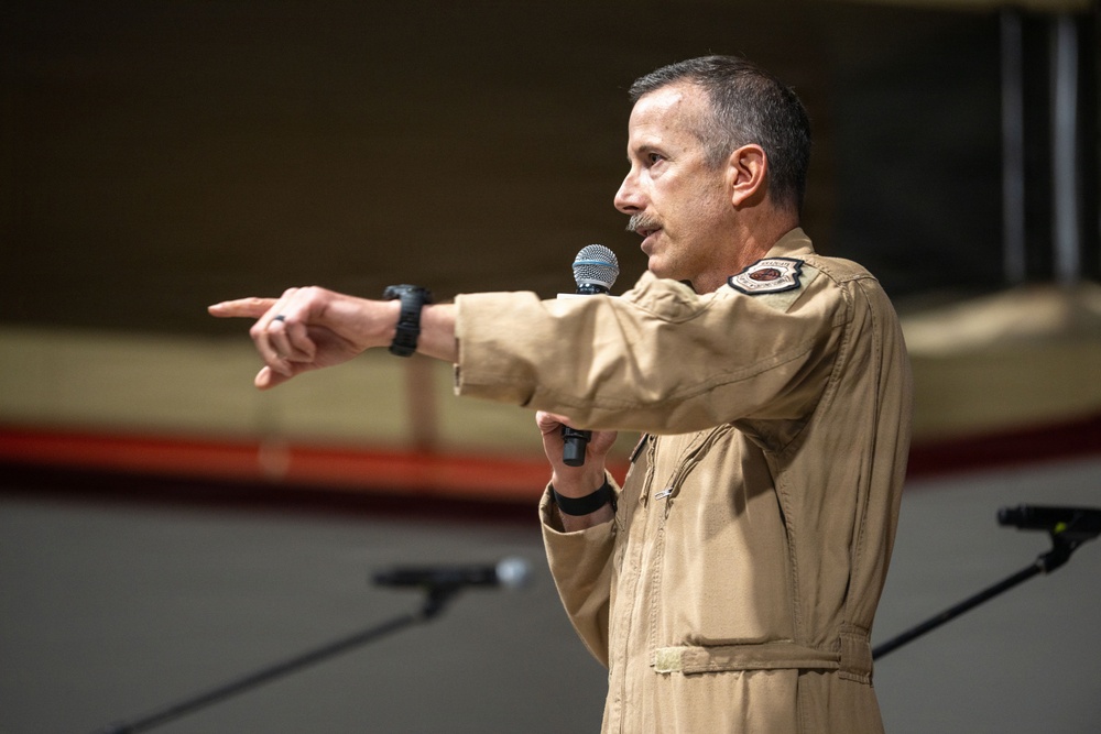A middle-aged man wearing a tan/khaki military flight suit is speaking into a handheld microphone while pointing with his left hand. He appears to be addressing an audience, likely in a hangar or large indoor military facility. A shoulder patch is visible on his right sleeve, indicating military affiliation.