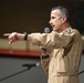 A middle-aged man wearing a tan/khaki military flight suit is speaking into a handheld microphone while pointing with his left hand. He appears to be addressing an audience, likely in a hangar or large indoor military facility. A shoulder patch is visible on his right sleeve, indicating military affiliation.