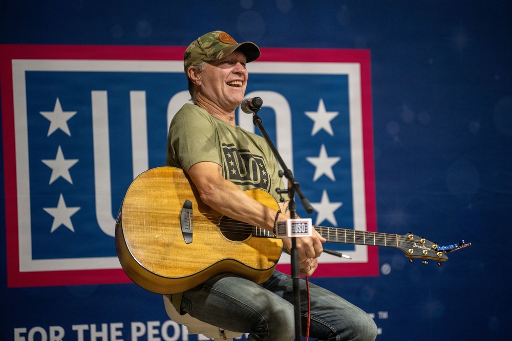 A middle-aged man is performing at a USO event, seated and playing an acoustic guitar while singing into a microphone. He is wearing a camouflage cap, an olive green USO t-shirt, and jeans, and appears to be smiling or laughing during the performance. The USO logo and tagline 'For the People' are prominently displayed on the backdrop behind him.