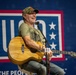 A middle-aged man is performing at a USO event, seated and playing an acoustic guitar while singing into a microphone. He is wearing a camouflage cap, an olive green USO t-shirt, and jeans, and appears to be smiling or laughing during the performance. The USO logo and tagline 'For the People' are prominently displayed on the backdrop behind him.