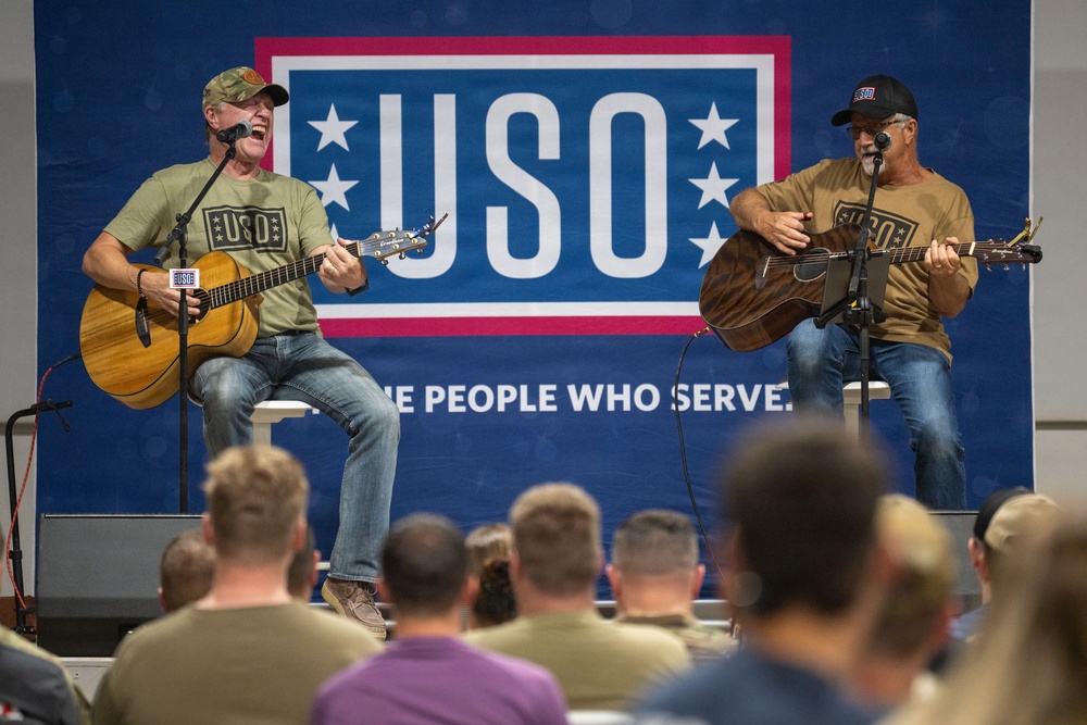 Two musicians are performing acoustic guitars on a stage at a USO event, both wearing matching olive/tan USO-branded t-shirts. They are seated on stools in front of a large blue USO banner reading 'THE PEOPLE WHO SERVE.' An audience of what appears to be military personnel watches from the foreground.