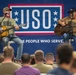 Two musicians are performing acoustic guitars on a stage at a USO event, both wearing matching olive/tan USO-branded t-shirts. They are seated on stools in front of a large blue USO banner reading 'THE PEOPLE WHO SERVE.' An audience of what appears to be military personnel watches from the foreground.