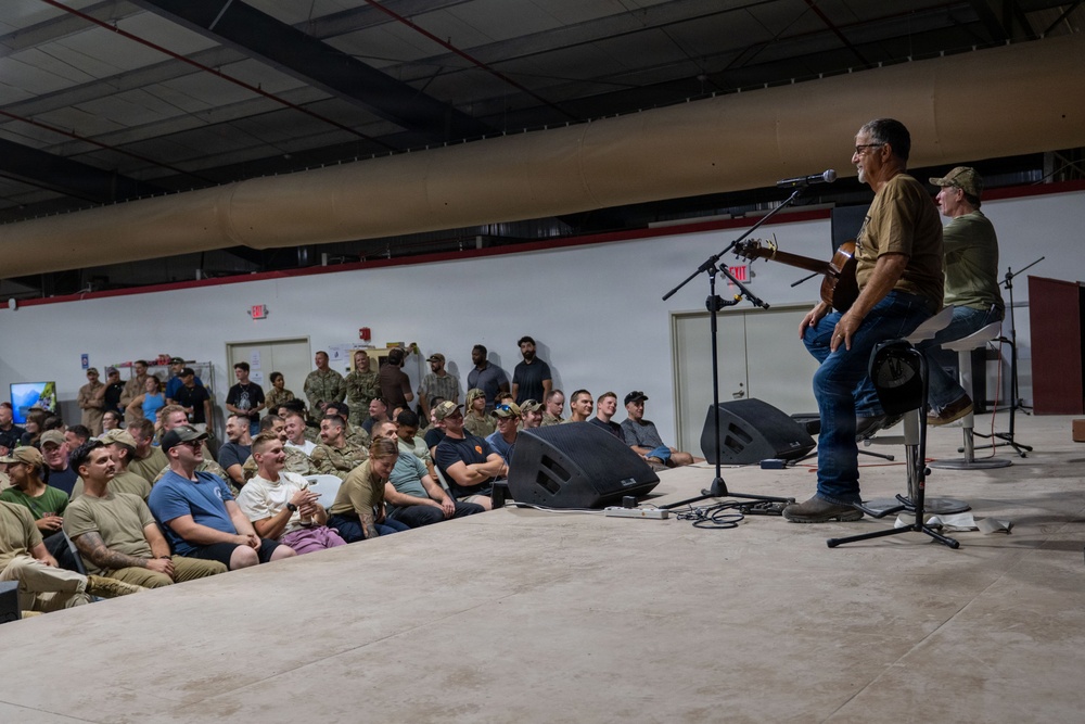 A live acoustic music performance is taking place inside what appears to be a military hangar or large indoor facility. Two performers are on a raised stage — one playing guitar and one seated — performing for a large audience of what appear to be military personnel and civilians. The crowd is engaged and visibly enjoying the performance, with many smiling and laughing.