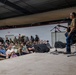A live acoustic music performance is taking place inside what appears to be a military hangar or large indoor facility. Two performers are on a raised stage — one playing guitar and one seated — performing for a large audience of what appear to be military personnel and civilians. The crowd is engaged and visibly enjoying the performance, with many smiling and laughing.