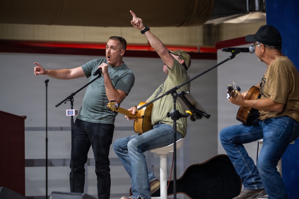 Three men are performing on a stage at what appears to be a USO event, identifiable by a USO-branded microphone stand. One man stands at the front singing into a handheld microphone while gesturing expressively, while two others sit behind him playing acoustic guitars. The setting appears to be an indoor venue such as a gymnasium or hangar.