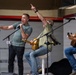 Three men are performing on a stage at what appears to be a USO event, identifiable by a USO-branded microphone stand. One man stands at the front singing into a handheld microphone while gesturing expressively, while two others sit behind him playing acoustic guitars. The setting appears to be an indoor venue such as a gymnasium or hangar.