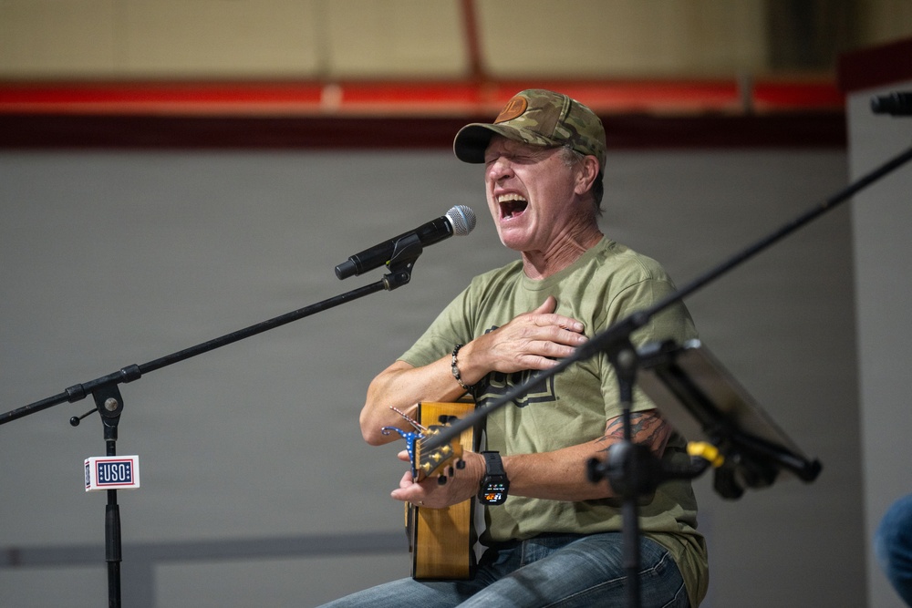 A middle-aged man is performing at a USO event, singing passionately into a microphone while holding an acoustic guitar. He is wearing a camouflage cap, olive green t-shirt, jeans, and has a tattoo visible on his left arm. The setting appears to be an indoor venue such as a gymnasium or hangar.