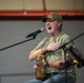 A middle-aged man is performing at a USO event, singing passionately into a microphone while holding an acoustic guitar. He is wearing a camouflage cap, olive green t-shirt, jeans, and has a tattoo visible on his left arm. The setting appears to be an indoor venue such as a gymnasium or hangar.