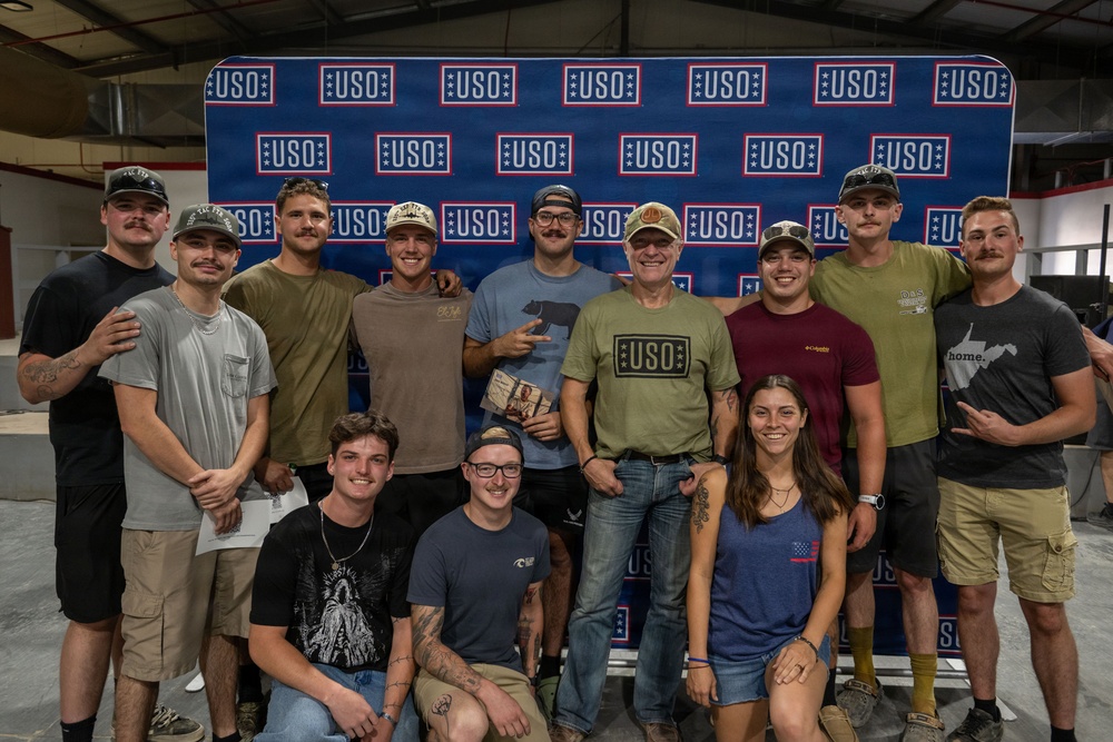 A group of approximately 13 people pose together in front of a USO branded step-and-repeat backdrop. The group includes men and women of varying ages, with one individual wearing a USO-branded t-shirt and holding what appears to be a gift or award item. The setting appears to be an indoor industrial or warehouse-type venue.