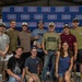 A group of approximately 13 people pose together in front of a USO branded step-and-repeat backdrop. The group includes men and women of varying ages, with one individual wearing a USO-branded t-shirt and holding what appears to be a gift or award item. The setting appears to be an indoor industrial or warehouse-type venue.