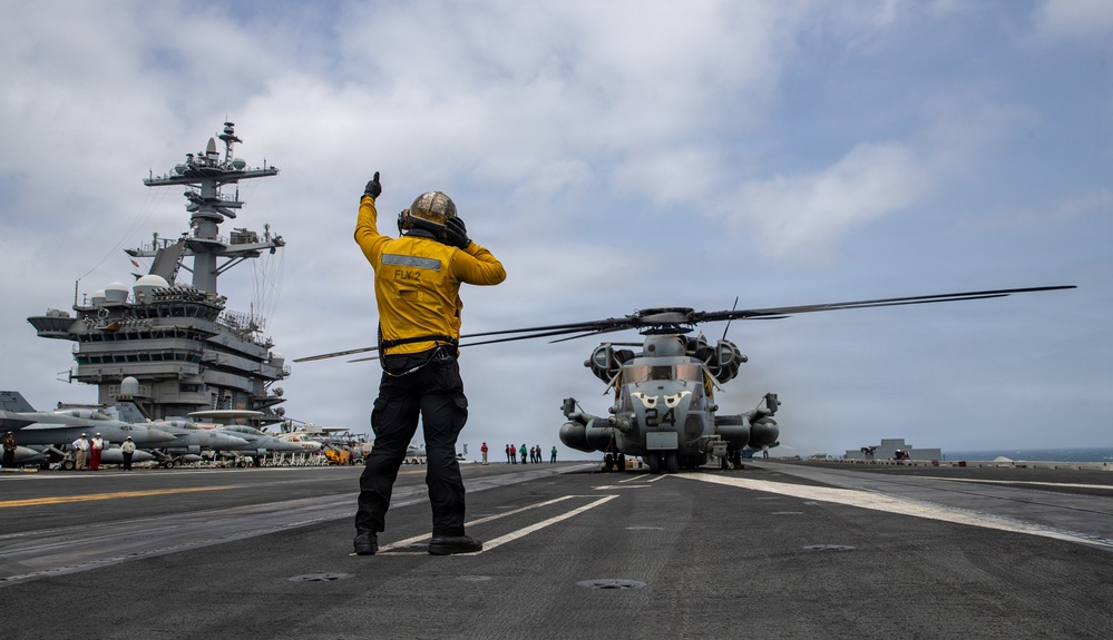 A flight deck crew member in a yellow jersey is directing a large military helicopter (numbered '24') on the flight deck of a U.S. Navy aircraft carrier. The crew member, identified by 'FLK 2' on their back, is giving hand signals while wearing a flight deck helmet and ear protection. Multiple fixed-wing aircraft and additional personnel are visible in the background near the carrier's island superstructure.