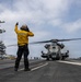 A flight deck crew member in a yellow jersey is directing a large military helicopter (numbered '24') on the flight deck of a U.S. Navy aircraft carrier. The crew member, identified by 'FLK 2' on their back, is giving hand signals while wearing a flight deck helmet and ear protection. Multiple fixed-wing aircraft and additional personnel are visible in the background near the carrier's island superstructure.
