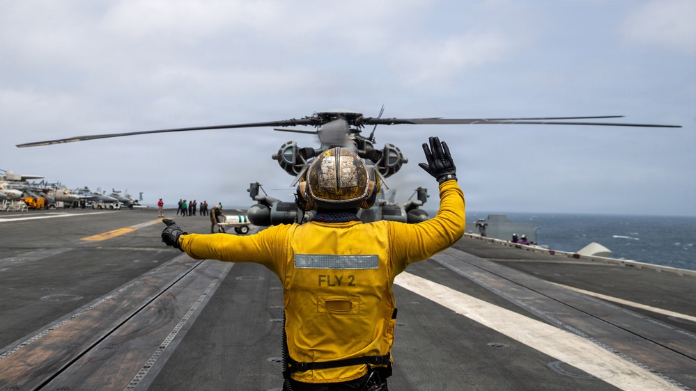 A flight deck crew member wearing a yellow jersey labeled 'FLY 2' signals to a military helicopter on the flight deck of an aircraft carrier at sea. The scene depicts standard carrier flight deck operations with multiple personnel and aircraft visible in the background. The overcast sky and open ocean are visible in the background, indicating active at-sea operations.