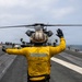 A flight deck crew member wearing a yellow jersey labeled 'FLY 2' signals to a military helicopter on the flight deck of an aircraft carrier at sea. The scene depicts standard carrier flight deck operations with multiple personnel and aircraft visible in the background. The overcast sky and open ocean are visible in the background, indicating active at-sea operations.