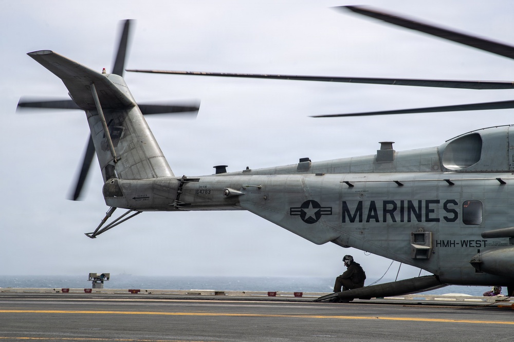 A U.S. Marines heavy-lift helicopter, identified as bureau number 164783 and assigned to HMH-WEST, is positioned on what appears to be the flight deck of a naval vessel. The helicopter's rotors are in motion, visible as motion blur in the frame. A crew member in flight gear is visible crouching near the rear of the aircraft.