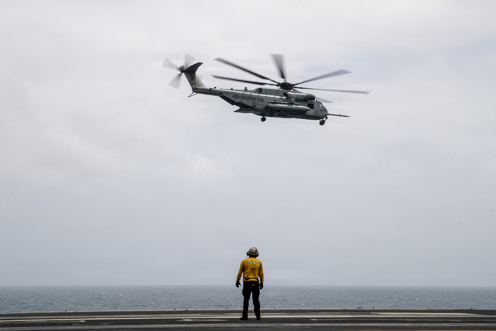 A U.S. Marines helicopter, identifiable as a CH-53E Super Stallion, hovers above the flight deck of a naval vessel over open ocean. A flight deck crew member wearing a yellow jersey and helmet stands below, likely serving as a landing signal officer or plane handler guiding the aircraft. The overcast sky and sea horizon are visible in the background.