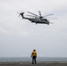 A U.S. Marines helicopter, identifiable as a CH-53E Super Stallion, hovers above the flight deck of a naval vessel over open ocean. A flight deck crew member wearing a yellow jersey and helmet stands below, likely serving as a landing signal officer or plane handler guiding the aircraft. The overcast sky and sea horizon are visible in the background.