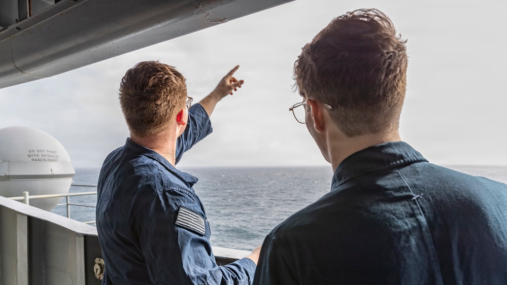 Two individuals stand on the deck of a vessel, looking out toward the open ocean. The person on the left, wearing a blue uniform with a subdued American flag patch on the sleeve, is pointing toward something in the distance. The person on the right, wearing glasses and a dark blue shirt, appears to be observing the direction being indicated.