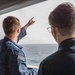 Two individuals stand on the deck of a vessel, looking out toward the open ocean. The person on the left, wearing a blue uniform with a subdued American flag patch on the sleeve, is pointing toward something in the distance. The person on the right, wearing glasses and a dark blue shirt, appears to be observing the direction being indicated.