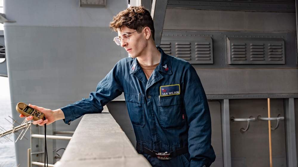 A young person wearing a navy blue military coverall uniform with a name tag reading 'IAN WILSON' is standing on what appears to be the deck or exterior walkway of a naval vessel. They are holding a tan/olive-colored handheld device, possibly a radio or GPS unit, while leaning against a railing. The background shows gray painted metal structures consistent with a military ship's superstructure.