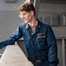 A young person wearing a navy blue military coverall uniform with a name tag reading 'IAN WILSON' is standing on what appears to be the deck or exterior walkway of a naval vessel. They are holding a tan/olive-colored handheld device, possibly a radio or GPS unit, while leaning against a railing. The background shows gray painted metal structures consistent with a military ship's superstructure.