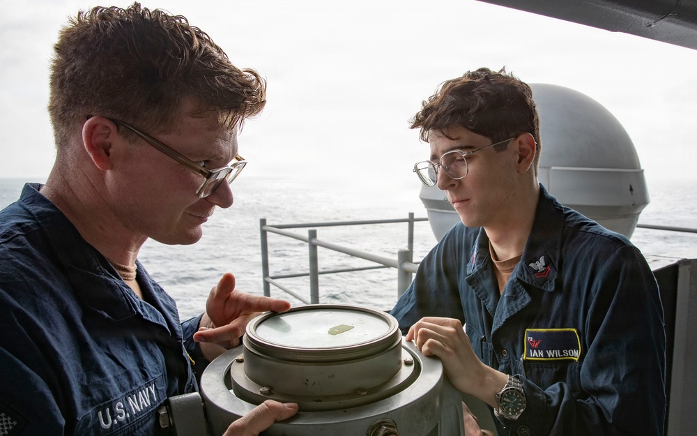 Two U.S. Navy personnel are examining a nautical compass or pelorus mounted on the deck of a naval vessel at sea. The individual on the right, identified by a name tag as 'Ian Wilson,' appears to be receiving instruction or guidance from the senior sailor on the left. Both are wearing standard Navy coveralls and glasses, engaged in what appears to be a navigation training exercise.