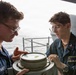 Two U.S. Navy personnel are examining a nautical compass or pelorus mounted on the deck of a naval vessel at sea. The individual on the right, identified by a name tag as 'Ian Wilson,' appears to be receiving instruction or guidance from the senior sailor on the left. Both are wearing standard Navy coveralls and glasses, engaged in what appears to be a navigation training exercise.