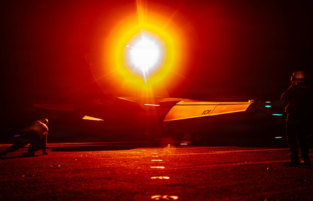 A night-time scene on what appears to be an aircraft carrier flight deck, with a military jet aircraft (marked '101') illuminated by an intense afterburner or engine exhaust flare. The extreme brightness of the engine exhaust creates a dramatic orange and white light bloom dominating the frame. At least two flight deck personnel in helmets are visible flanking the aircraft.