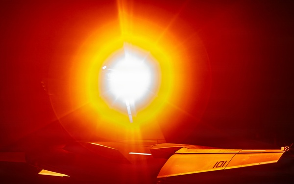 A night-time scene on what appears to be an aircraft carrier flight deck, with a military jet aircraft (marked '101') illuminated by an intense afterburner or engine exhaust flare. The extreme brightness of the engine exhaust creates a dramatic orange and white light bloom dominating the frame. At least two flight deck personnel in helmets are visible flanking the aircraft.