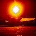 A night-time scene on what appears to be an aircraft carrier flight deck, with a military jet aircraft (marked '101') illuminated by an intense afterburner or engine exhaust flare. The extreme brightness of the engine exhaust creates a dramatic orange and white light bloom dominating the frame. At least two flight deck personnel in helmets are visible flanking the aircraft.