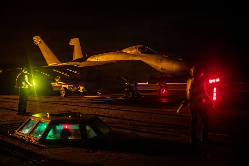 A military jet aircraft, identifiable as an F/A-18 Hornet or Super Hornet with markings 'VFA-195' and number '405', is photographed on what appears to be an aircraft carrier flight deck at night. The scene is illuminated by warm amber/yellow and red lighting, with deck crew personnel visible on both sides of the aircraft. Illuminated deck hatches and signal wands are visible in the foreground.