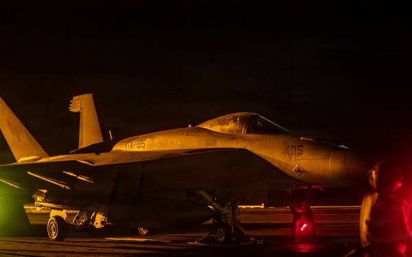 A military jet aircraft, identifiable as an F/A-18 Hornet or Super Hornet with markings 'VFA-195' and number '405', is photographed on what appears to be an aircraft carrier flight deck at night. The scene is illuminated by warm amber/yellow and red lighting, with deck crew personnel visible on both sides of the aircraft. Illuminated deck hatches and signal wands are visible in the foreground.