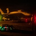 A military jet aircraft, identifiable as an F/A-18 Hornet or Super Hornet with markings 'VFA-195' and number '405', is photographed on what appears to be an aircraft carrier flight deck at night. The scene is illuminated by warm amber/yellow and red lighting, with deck crew personnel visible on both sides of the aircraft. Illuminated deck hatches and signal wands are visible in the foreground.