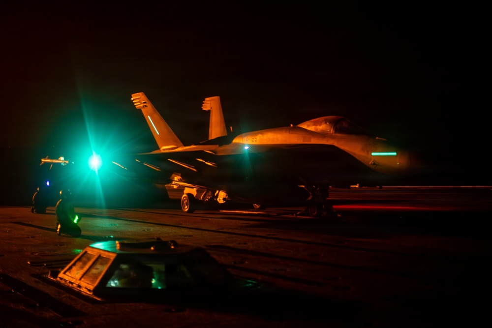 A military jet aircraft, consistent with an F/A-18 Hornet or Super Hornet, is photographed on what appears to be an aircraft carrier flight deck during nighttime operations. The scene is illuminated by orange and teal/green artificial lighting, with ground crew personnel visible nearby. The low-light conditions and deck markings suggest active carrier flight deck operations.