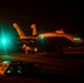 A military jet aircraft, consistent with an F/A-18 Hornet or Super Hornet, is photographed on what appears to be an aircraft carrier flight deck during nighttime operations. The scene is illuminated by orange and teal/green artificial lighting, with ground crew personnel visible nearby. The low-light conditions and deck markings suggest active carrier flight deck operations.
