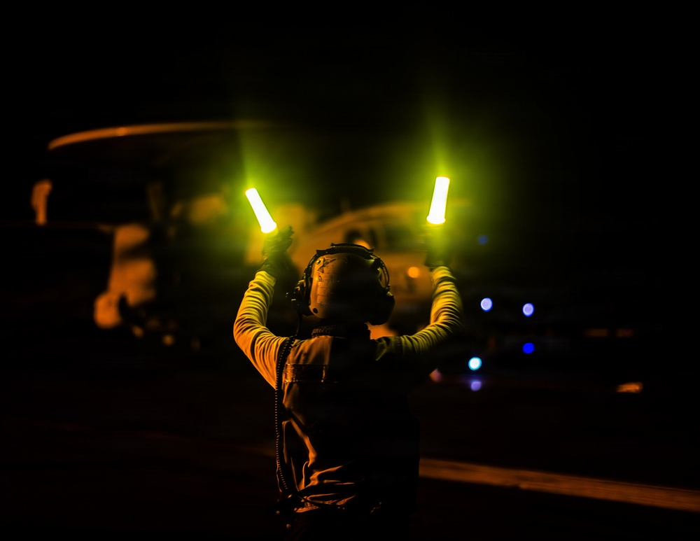 A military flight deck crew member is conducting nighttime aircraft marshalling operations, holding two illuminated wands (batons) raised above their head. The scene appears to take place on a ship's flight deck or airfield at night, with a large rotary-wing aircraft visible in the background. The image is dramatically lit by the glow of the marshalling wands and ambient aircraft lighting.
