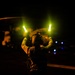 A military flight deck crew member is conducting nighttime aircraft marshalling operations, holding two illuminated wands (batons) raised above their head. The scene appears to take place on a ship's flight deck or airfield at night, with a large rotary-wing aircraft visible in the background. The image is dramatically lit by the glow of the marshalling wands and ambient aircraft lighting.