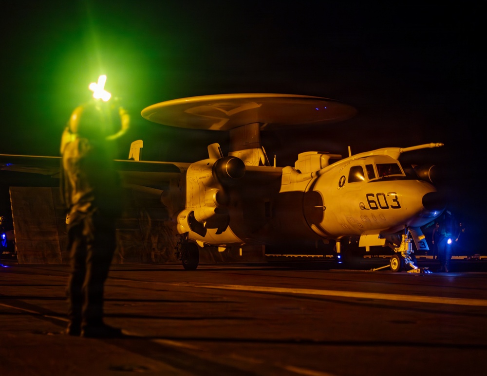 A night operations scene on what appears to be a naval aircraft carrier flight deck, showing a Grumman E-2 Hawkeye airborne early warning aircraft marked '603'. A flight deck crew member is visible in the foreground holding illuminated wands, with green and orange lighting dominating the scene. Additional personnel with blue lights are visible near the aircraft's landing gear.