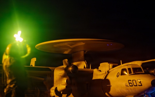 A night operations scene on what appears to be a naval aircraft carrier flight deck, showing a Grumman E-2 Hawkeye airborne early warning aircraft marked '603'. A flight deck crew member is visible in the foreground holding illuminated wands, with green and orange lighting dominating the scene. Additional personnel with blue lights are visible near the aircraft's landing gear.