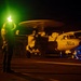 A night operations scene on what appears to be a naval aircraft carrier flight deck, showing a Grumman E-2 Hawkeye airborne early warning aircraft marked '603'. A flight deck crew member is visible in the foreground holding illuminated wands, with green and orange lighting dominating the scene. Additional personnel with blue lights are visible near the aircraft's landing gear.