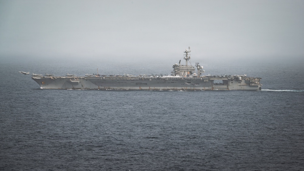 A large U.S. Navy aircraft carrier bearing hull number '73' is underway at sea under overcast, hazy conditions. The flight deck is populated with multiple aircraft, and at least one aircraft appears to be launching or departing from the bow. The vessel is moving through calm, dark ocean waters with a visible wake at the stern.