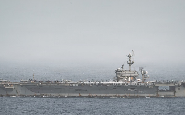 A large U.S. Navy aircraft carrier bearing hull number '73' is underway at sea under overcast, hazy conditions. The flight deck is populated with multiple aircraft, and at least one aircraft appears to be launching or departing from the bow. The vessel is moving through calm, dark ocean waters with a visible wake at the stern.